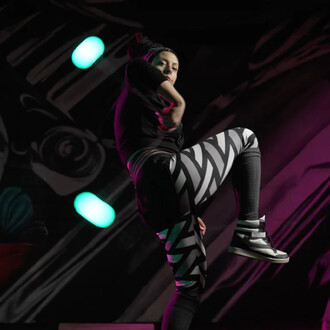 Personne en mouvement, exécutant une figure de jonglerie avec des bolas Coral Flowtoys, vêtue d'un haut noir et de leggings à motifs en noir et blanc. L'arrière-plan présente des lumières colorées dans des teintes vertes et violettes.