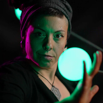Une personne avec des piercings et un chapeau noir se tient devant un fond sombre, illuminée par des lumières vertes et roses. Elle regarde la caméra avec une expression concentrée et maîtrise des bolas colorés dans ses mains.