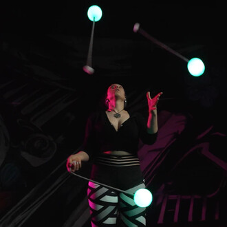 Une personne jongle avec des bolas lumineuses vertes dans une atmosphère sombre, mettant en valeur les éléments fluos. Elle est vêtue d’un haut noir et de leggings à motifs. L'arrière-plan présente une illustration stylisée.