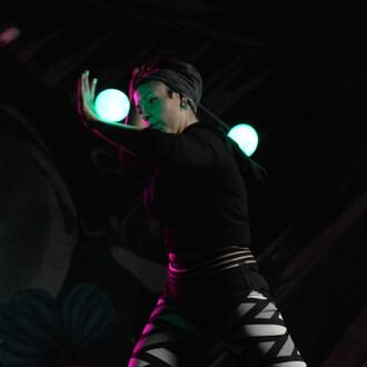 Femme en train de jongler avec des bolas, portant un haut noir et un pantalon à motif géométrique, dans un environnement sombre avec des lumières vertes en arrière-plan. Elle montre une posture concentrée et dynamique.