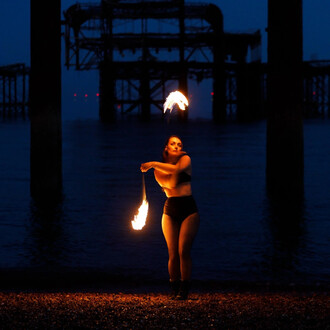 Une jongleuse exécute une performance avec des bolas enflammées sur une plage nocturne. En arrière-plan, on aperçoit des structures industrielles obscures. La jongleuse, habillée de façon minimaliste, est au centre de l'image, tandis que les flammes brill