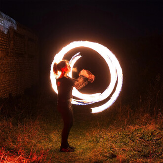 Une personne jonglant avec des bolas enflammées dans un environnement nocturne. Des cercles de feu brillants se déplacent autour d'elle, illuminant la scène sombre avec des reflets vifs. Le jardin en arrière-plan est peu éclairé et l'atmosphère est dynami