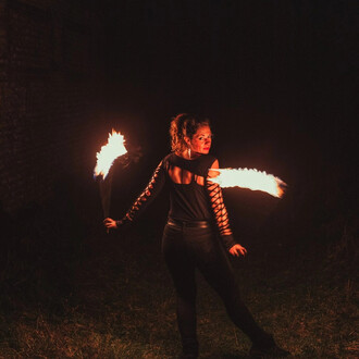 Femme en train de jongler avec des bolas enflammés, vêtue d'un haut noir à manches lacées, dans un environnement sombre. Les flammes illuminent son visage et créent une ambiance dynamique et spectaculaire.