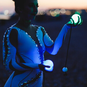 Jongleuse en tenue scintillante tenant des bolas LED Echo Orbit, avec des sphères lumineuses dans chaque main, au crépuscule. Les bolas émettent des lumières aux teintes bleu et vert, tandis que l'arrière-plan montre des lumières floues évoquant une ambia