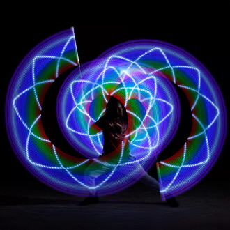 Personne jonglant avec des bolas lumineuses, créant des motifs colorés en mouvement, sur un fond sombre. Les lumières des bolas forment des formes géométriques éclatantes.