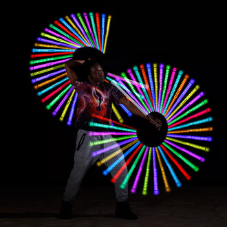 Un jongleur exécute des mouvements avec des bolas lumineuses, créant des spirales colorées de lumière dans un fond noir. Il porte un t-shirt aux motifs galactiques et un pantalon de sport. Les bolas émettent des lumières vives dans plusieurs couleurs, met