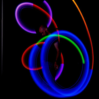 Bolas lumineuses en mouvement éclairant un fond noir, avec des tracés de lumière bleu, vert, orange, rouge et violet, créant des formes dynamiques. Un jongleur est partiellement visible, concentré sur sa performance.