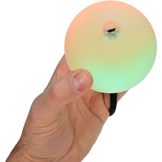 Bola lumineuse Supernova tenue dans une main, présentant un design sphérique avec des couleurs dégradées allant de l'orange au vert. On peut voir la prise de chargement sur le dessus de la bola.