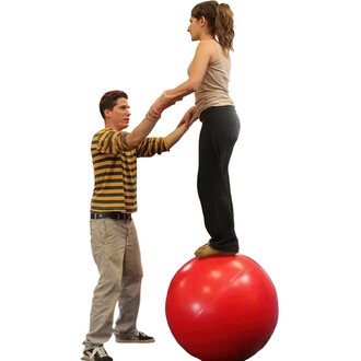 Un homme en t-shirt rayé tient les mains d'une femme qui se tient en équilibre sur une grande boule rouge de 70 cm. La femme est en position debout, les jambes légèrement fléchies, tandis que l'homme est en dessous pour la soutenir, montrant une dynamique