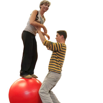 Deux personnes sont en train de jongler avec une boule d'équilibre rouge de 70 cm. Une femme se tient debout sur la boule, tandis qu'un homme la soutient en tenant ses mains. Ils sont tous les deux en vêtements décontractés, dans un environnement clair.