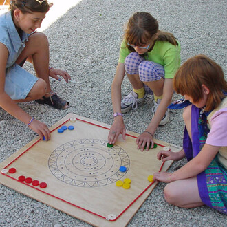 Trois enfants jouent à un jeu de société sur une surface en gravier. Ils utilisent un plateau en bois avec un motif circulaire et des marques numérotées. Les enfants placent des jetons colorés de différentes couleurs (bleu, vert, rouge et jaune) sur le pl