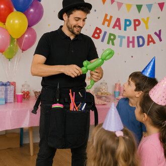 Animateur d'anniversaire portant une ceinture tablier noire remplie d'outils pour la sculpture de ballons, en train de modeler un ballon vert en forme d'animal devant des enfants lors d'une fête décorée de ballons et d'un 'Happy Birthday'.