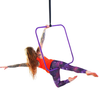 Femme en position acrobatique, suspendue à un cerceau aérien carré de couleur violet, portant un débardeur orange et un legging à motif galactique. Elle s'étire avec un bras tendu et l'autre tenant le cerceau, les pieds nus. Le fond est blanc.