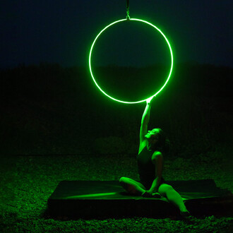 Artiste assise sous un cerceau aérien lumineux vert, dans un cadre extérieur nocturne.