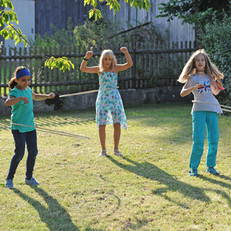 Trois enfants s'amusent dans un jardin en utilisant des bâtons de jonglerie. L'un d'eux porte une robe à fleurs, un autre une t-shirt gris avec un drapeau britannique, et le dernier est vêtu d'un haut turquoise. Ils montrent des expressions de joie tout e