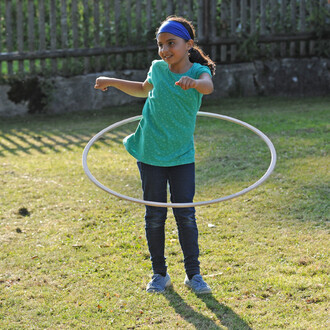 Enfant souriant en train de jongler avec un hula hoop dans un jardin, portant un t-shirt vert à pois et un bandeau bleu.