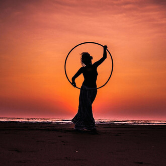 Silhouette d'une femme tenant un cerceau de hula hoop au coucher du soleil, avec l'océan en arrière-plan et des couleurs orange et violet dans le ciel.