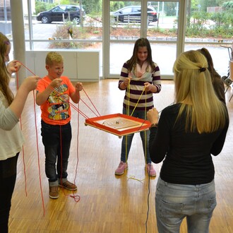 Group of children engaging in a team-building activity with the Pedalo® 3 game. They are holding colorful strings attached to a circular wooden platform, concentrating on coordinating their movements together. The setting is an indoor space with large win