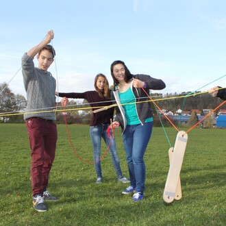 Quatre jeunes personnes interagissent dans un espace vert, en utilisant des cordes liées à un appareil Pedalo. Deux garçons et deux filles semblent coopérer pour manipuler les cordes colorées. L'ambiance est dynamique et ludique, avec un ciel dégagé en ar