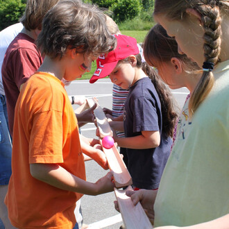 Un groupe d'enfants participent à une activité extérieure, s'entraidant pour maintenir une balle rose sur des planches en bois alignées. L'un des enfants, portant un t-shirt orange, se concentre sur l'équilibre de la balle, tandis qu'une fille en casquett