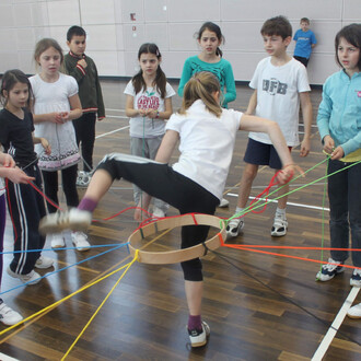 Groupe d'enfants en train de jouer ensemble dans une salle, autour d'un cercle fait de fil coloré. Une fille s'apprête à passer à travers le cercle en levant une jambe. Les autres enfants, observe avec intérêt, certains tiennent des fils d'autres couleurs