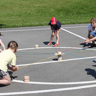 Des enfants et un adulte jouent ensemble sur un terrain extérieur, avec quatre poteaux en bois placés au centre. Ils utilisent des cordes de différentes couleurs, tirant chacune dans une direction. L'ambiance est conviviale et ensoleillée, avec de l'herbe