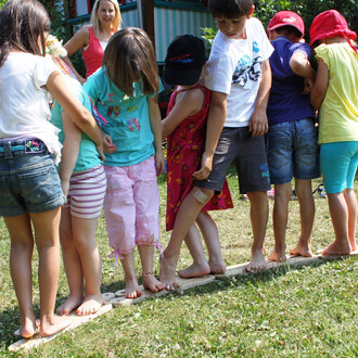 Un groupe d'enfants pieds nus se tient en ligne sur une planche de bois posée sur l'herbe. Ils sont en train de jouer ensemble, portant des vêtements colorés et montrant des expressions de joie. Une adulte est en arrière-plan, observant et souriant.