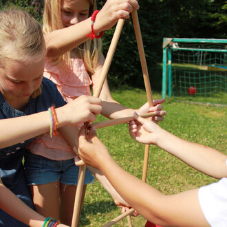 Des enfants s'amusent à construire une structure en utilisant des bâtons en bois. Ils sont concentrés et travaillent ensemble, les mains se rejoignant pour assembler les pièces sous un ciel ensoleillé, avec un fond de jardin verdoyant et un but de footbal