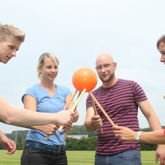 Quatre personnes s'affrontent en équipe pour faire tenir un ballon orange sur des bâtons. Ils se tiennent debout sur une pelouse verte, sous un ciel clair. Les participants semblent concentrés et engagés dans le jeu.