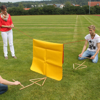 Un groupe de trois personnes est en train de jouer avec un équipement de jeu d'équipe sur un terrain vert. Une femme tient une corde attachée à un grand panneau jaune, tandis que deux hommes, assis au sol, manipulent des bâtons en bois de forme triangulai