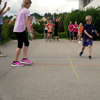 Des enfants s'amusent à une activité en plein air, avec un garçon en t-shirt bleu courant sur une surface en béton. Une fille portant un t-shirt violet se tient penchée près de lui. Des camarades en arrière-plan applaudissent et encouragent. Des balles ja