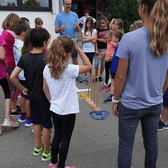 Groupe d'enfants rassemblés autour d'un adulte qui explique une activité. Au centre, plusieurs bâtons tenus debout, chacun portant un œuf. Des cerceaux de différentes couleurs sont disposés au sol, tandis que les enfants, souriants, observent attentivemen