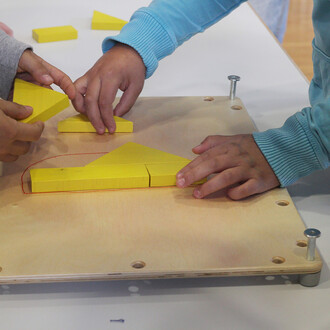 Mains d'enfants manipulant des pièces en bois jaunes sur une table en bois, dans le cadre d'un jeu de groupe. Les pièces incluent des formes géométriques, comme des triangles et des blocs.