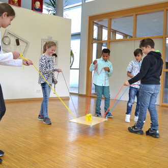 Groupe d'enfants jouant avec le jeu d'équipe Pedalo® 3, utilisant des cordes de différentes couleurs pour tirer sur une plateforme en bois au sol, dans une salle lumineuse.