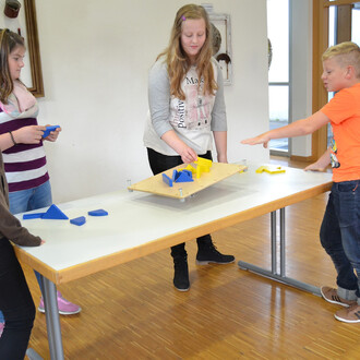 Quatre enfants sont rassemblés autour d'une table. Au centre, une planche supporte des pièces en bois de couleur jaune et bleue, disposées de manière créative. Deux enfants, une fille aux cheveux longs et une autre avec un foulard, manipulent des pièces b