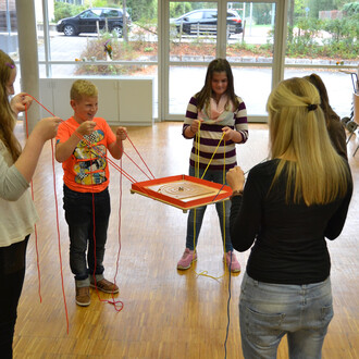 Group of children engaging in a team-building activity with the Pedalo® 3 game. They are holding colorful strings attached to a circular wooden platform, concentrating on coordinating their movements together. The setting is an indoor space with large win