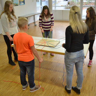 Cinq enfants sont réunis autour d'une table avec un jeu de collaboration. Ils tiennent des cordes de différentes couleurs reliées à une structure en bois posée sur la table. L'ambiance est dynamique et ludique, avec un sol en bois et des murs clairs en ar