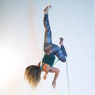 Femme suspendue à une corde lisse, en position acrobatique, portant un haut et un legging aux teintes sombres, cheveux longs et ondulés. Fond clair et lumineux.