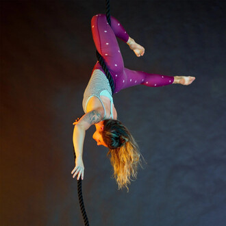 Femme suspendue à une corde lisse, en position d'acrobatie, portant un justaucorps rayé et un legging violet orné d'étoiles. Fond sombre avec un éclairage coloré.