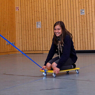 Fille souriante assise sur un chariot jaune à roulettes, tirée par une corde bleue dans une salle aux murs en bois.