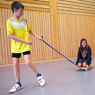 Un garçon portant un t-shirt jaune et un short noir tire une planche à roulettes avec une corde bleue, sur laquelle une fille souriante est assise. Ils évoluent dans une salle avec des murs en bois et un sol gris.