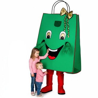 Mascotte de sac cadeau en forme de grand sac vert avec un sourire, des yeux ronds et un nœud doré. Deux enfants, l'un debout et l'autre plus petit, interagissent avec la mascotte, tous deux portant des pulls roses. Fond blanc.