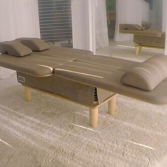 Table de massage moderne avec deux coussins, finition marron, placée sur un tapis clair. L'environnement est lumineux avec des murs clairs et des reflets dans un miroir.