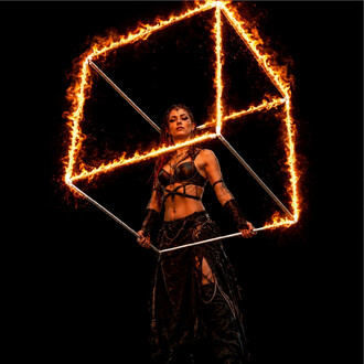Ein Feuerkünstler steht in einem geometrischen Würfel in Flammen; der Metallrahmen ist von orangefarbenen Flammen umgeben, vor schwarzem Hintergrund.