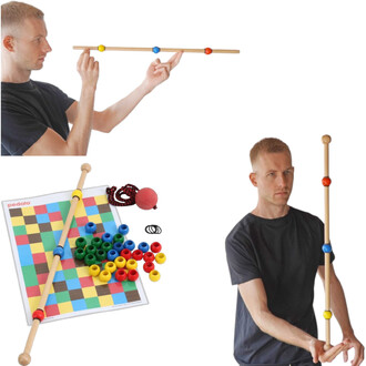 Un homme en t-shirt noir manipule une canne en bois avec des billes colorées (jaune, bleu, rouge) à chaque extrémité. En bas à gauche, un plateau de jeux avec un motif coloré, une canne similaire, des anneaux, une balle rouge et plusieurs pièces de différ