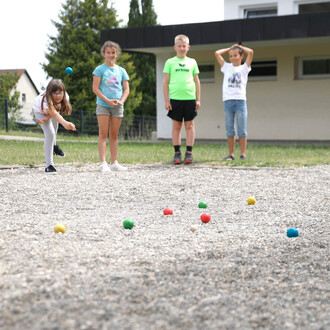 Quatre enfants jouent à un jeu de boules sur un terrain de graviers. Une fille s'apprête à lancer une boule bleue tandis que les autres l'observent. Les boules de différentes couleurs sont dispersées sur le sol. En arrière-plan, on voit une maison et des 