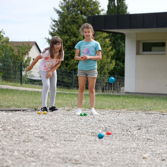 Deux filles jouent à un jeu de boules sur un terrain en gravier. L'une des filles, portant un t-shirt bleu, est en train de lancer une boule bleue, tandis que l'autre, portant un t-shirt coloré, observe avec un sourire. Plusieurs boules colorées sont disp