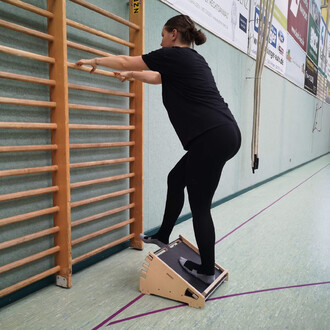 Femme en train de s'exercer sur une planche inclinée en bois, se tenant à une barre de gymnastique. Elle porte un t-shirt noir et des leggings noirs, avec des chaussettes. L'environnement est une salle de sport, avec un sol en vinyle et des murs de couleu