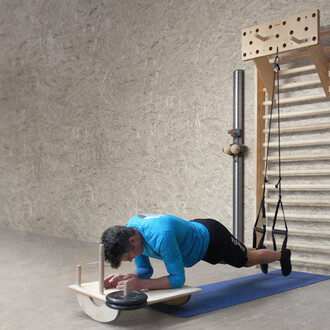 Un homme effectue un exercice de gainage sur une planche d'équilibre, avec des poids placés à une extrémité. Il porte un t-shirt bleu et un short noir. En arrière-plan, on peut voir un mur avec des éléments d'équipement de fitness, y compris une barre ver