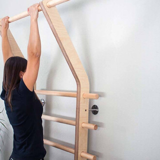 Femme en train de faire des tractions sur un appareil de musculation en bois, fixé au mur. L'appareil, de couleur claire, comprend plusieurs barres horizontales et verticales, offrant un support pour différents exercices. Le fond est sobre, avec un mur bl